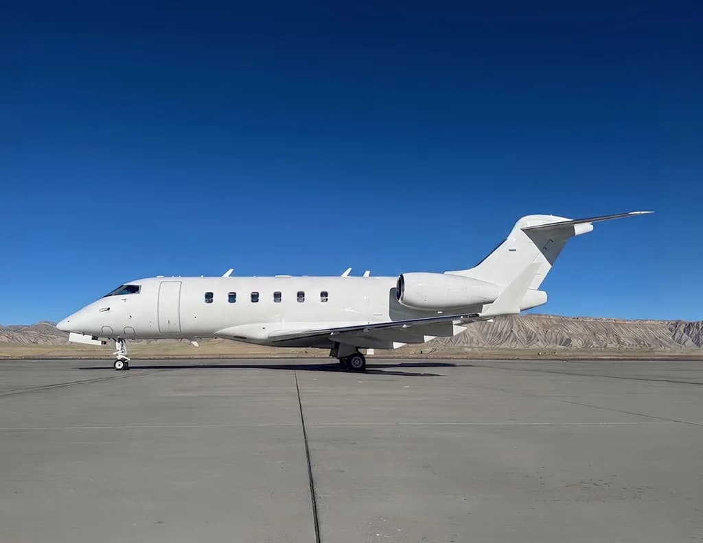 A white Bombardier Challenger 605 business jet parked on an airfield with mountains in the background.