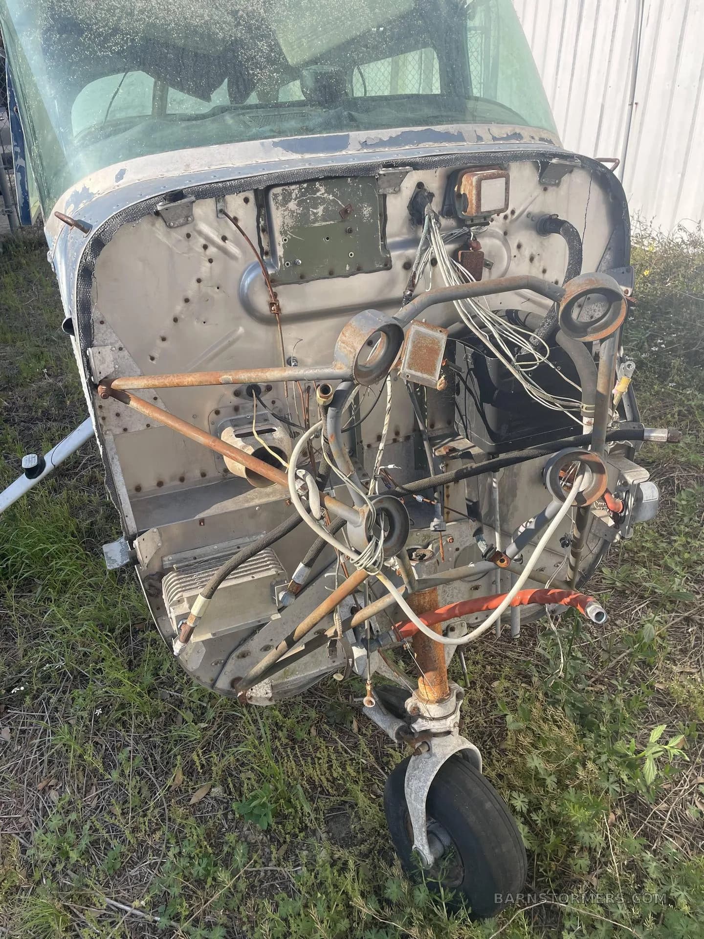A front view of a disassembled, weathered light aircraft fuselage, showing exposed engine bay components and the nose landing gear on grass.