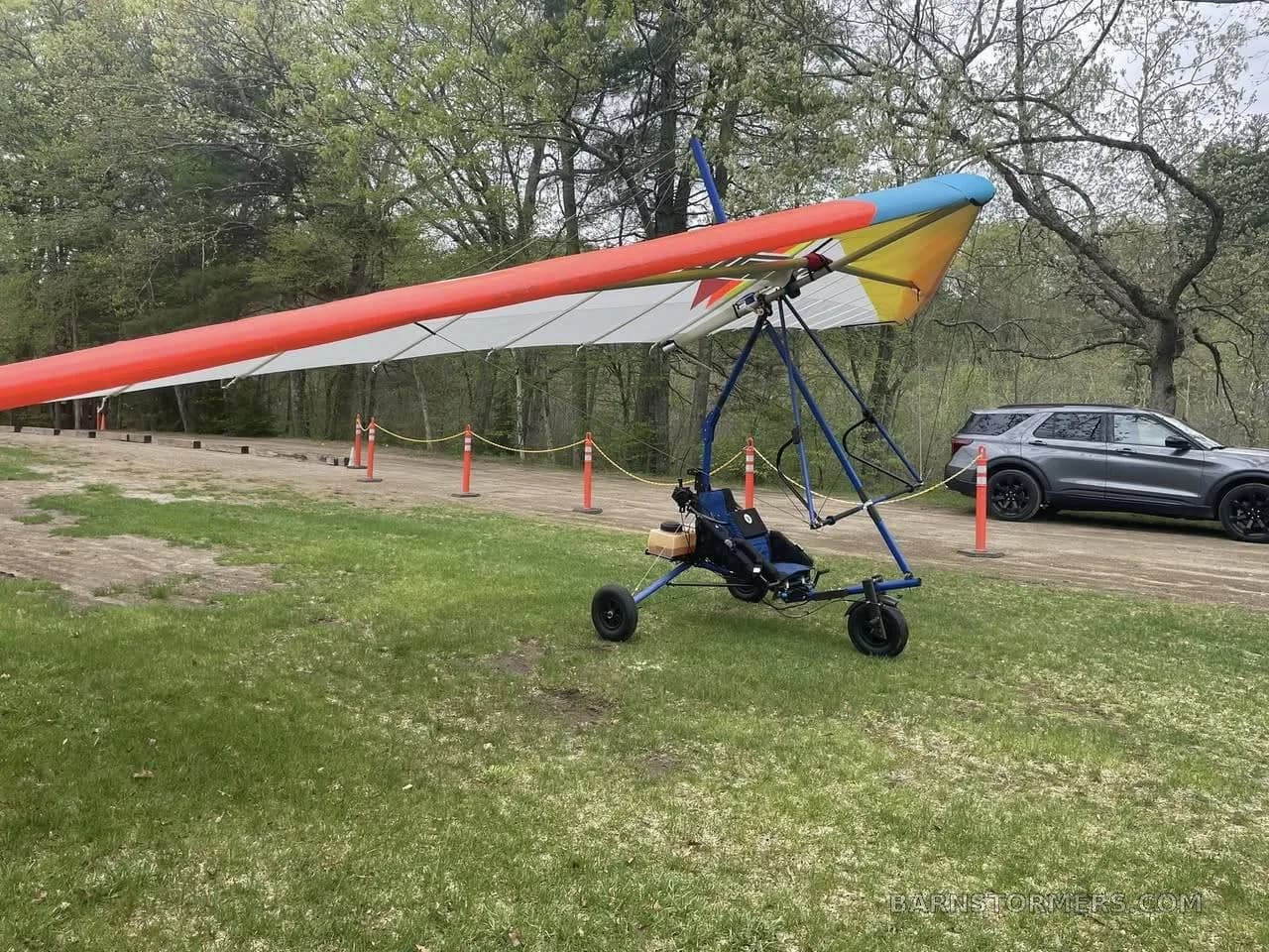 A powered hang glider with an orange, yellow, and white wing sits on a grassy patch next to a dirt path and a parked SUV.