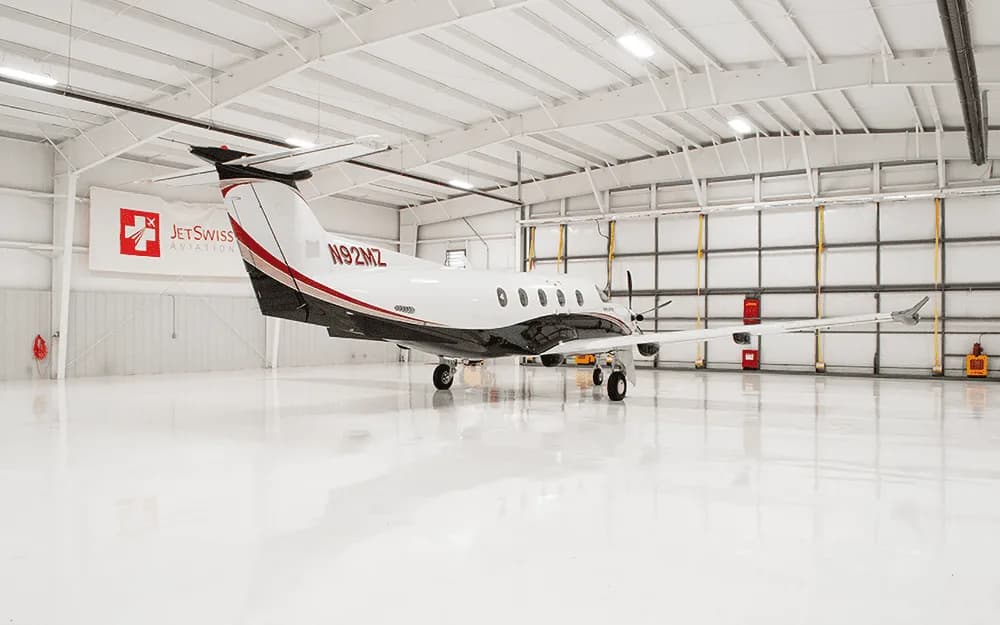 A white and black Pilatus PC-12 aircraft with tail number N92MZ is parked in a brightly lit white hangar, viewed from the rear-right.