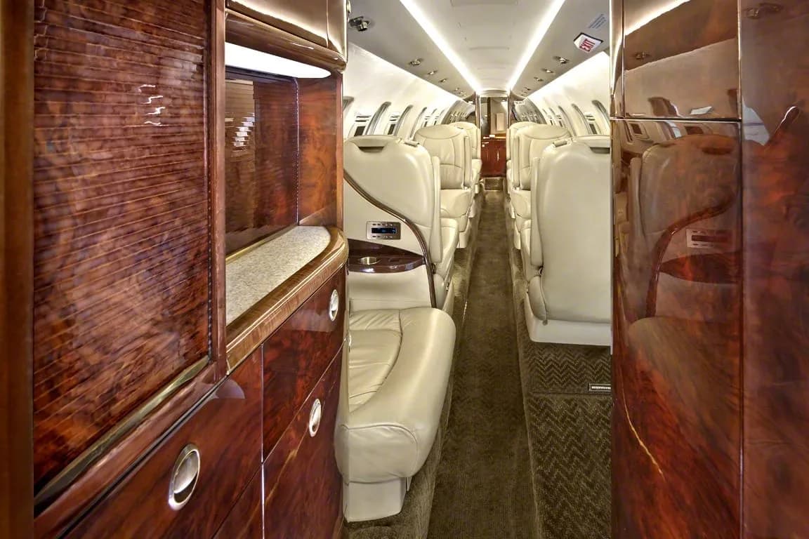 The long interior cabin of a private jet with beige leather seats, dark wood paneling, and overhead lighting.
