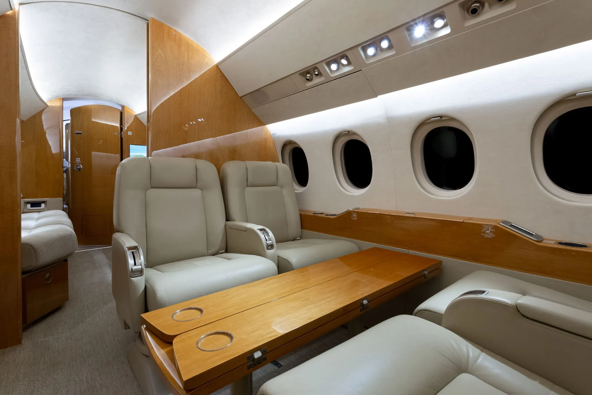 A view inside a private jet cabin showing two beige leather seats facing a wooden table, with a lounge area visible further back.