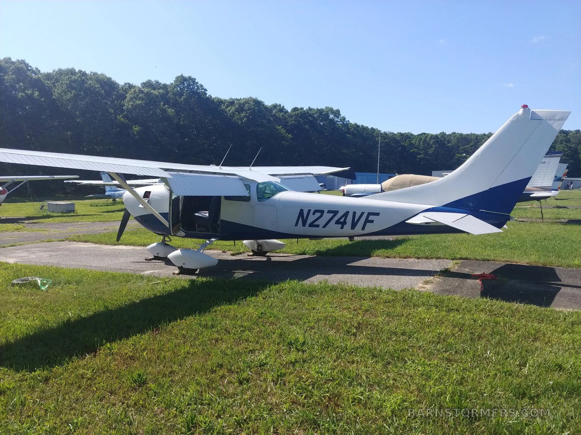 Side view of a white and blue single-engine aircraft (N274VF) with its door open, parked on an asphalt path next to grass.