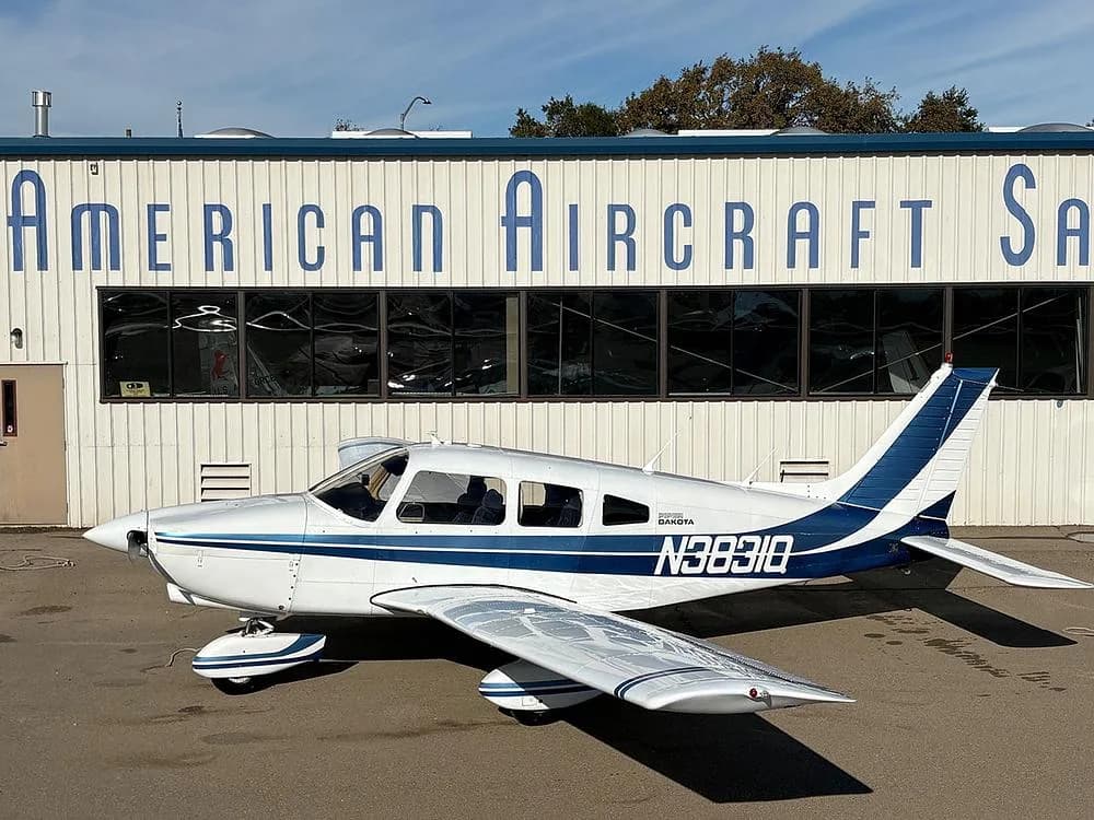 A white Piper Dakota aircraft with blue stripes, registration N3831Q, parked in front of a building labeled "American Aircraft Sales".