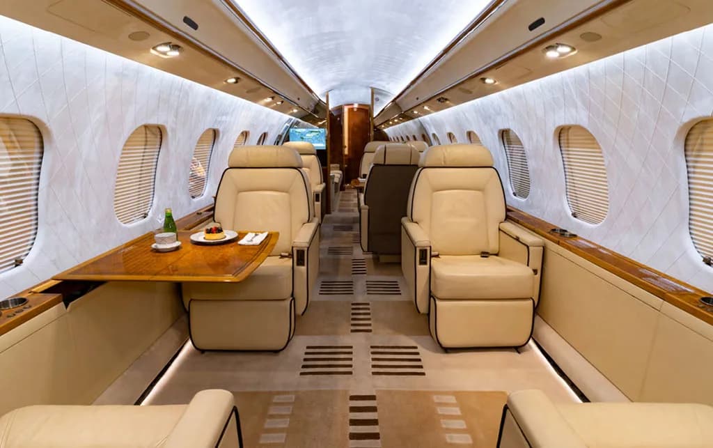 The luxurious interior cabin of a private jet with beige leather seats and a wooden table set with food.