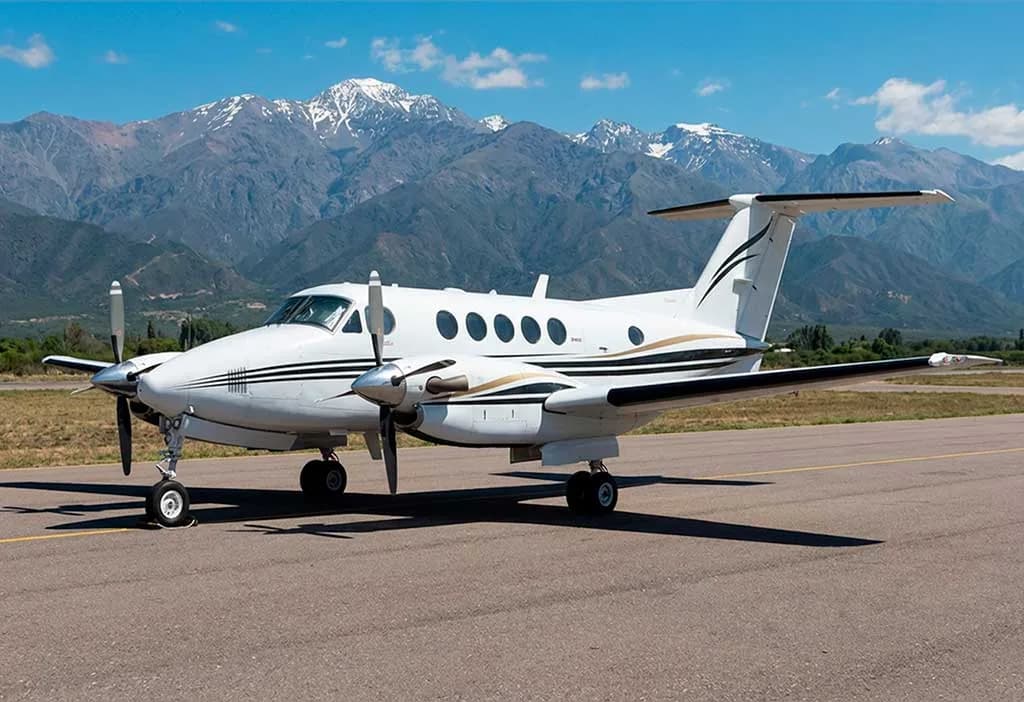 A white twin-turboprop aircraft with black and gold stripes is parked on an airfield with snow-capped mountains in the background.