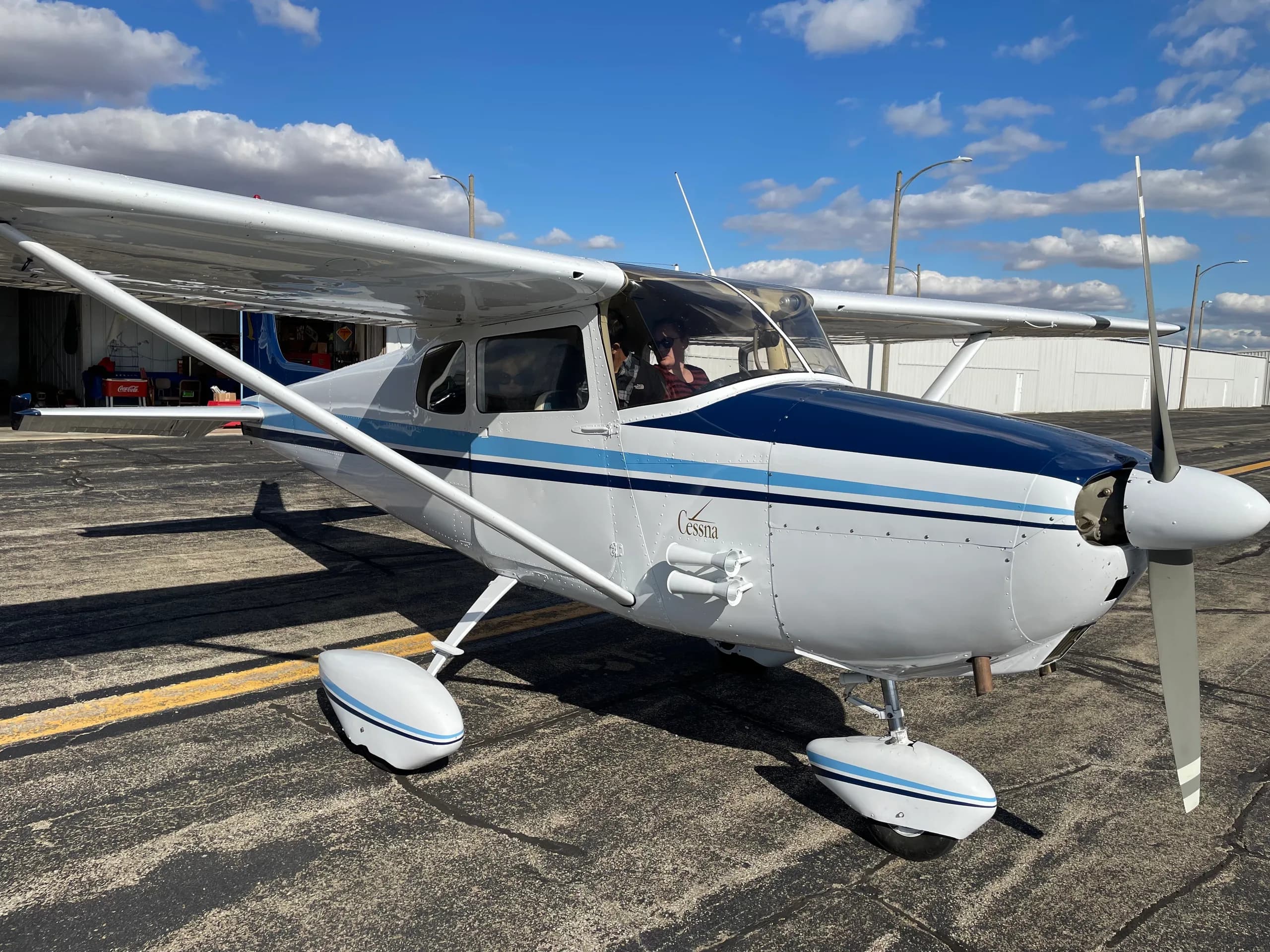 A white Cessna aircraft with blue stripes on the fuselage and wheel fairings, parked on an asphalt tarmac under a blue sky.