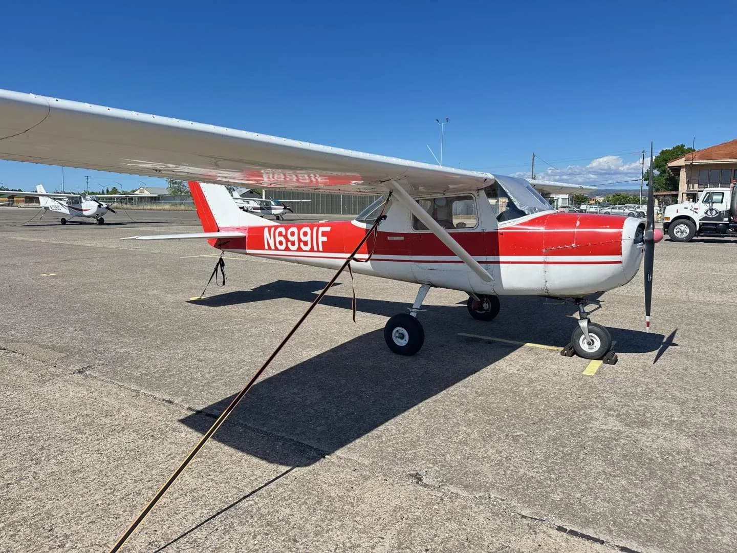A red and white Cessna 150, tail number N6991F, parked on an asphalt apron with other aircraft and buildings in the distance.