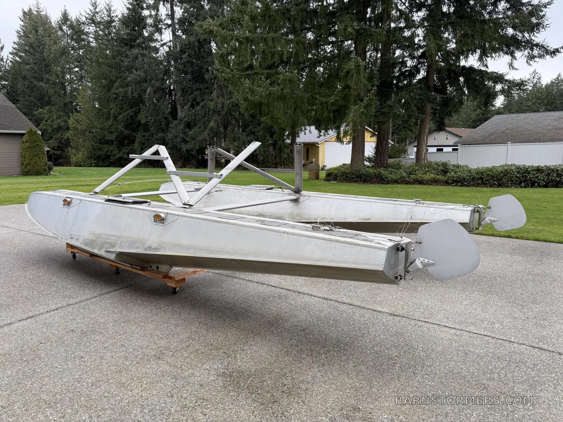 A pair of aluminum aircraft floats with mounting struts and rudders, viewed from the rear, on a concrete driveway.