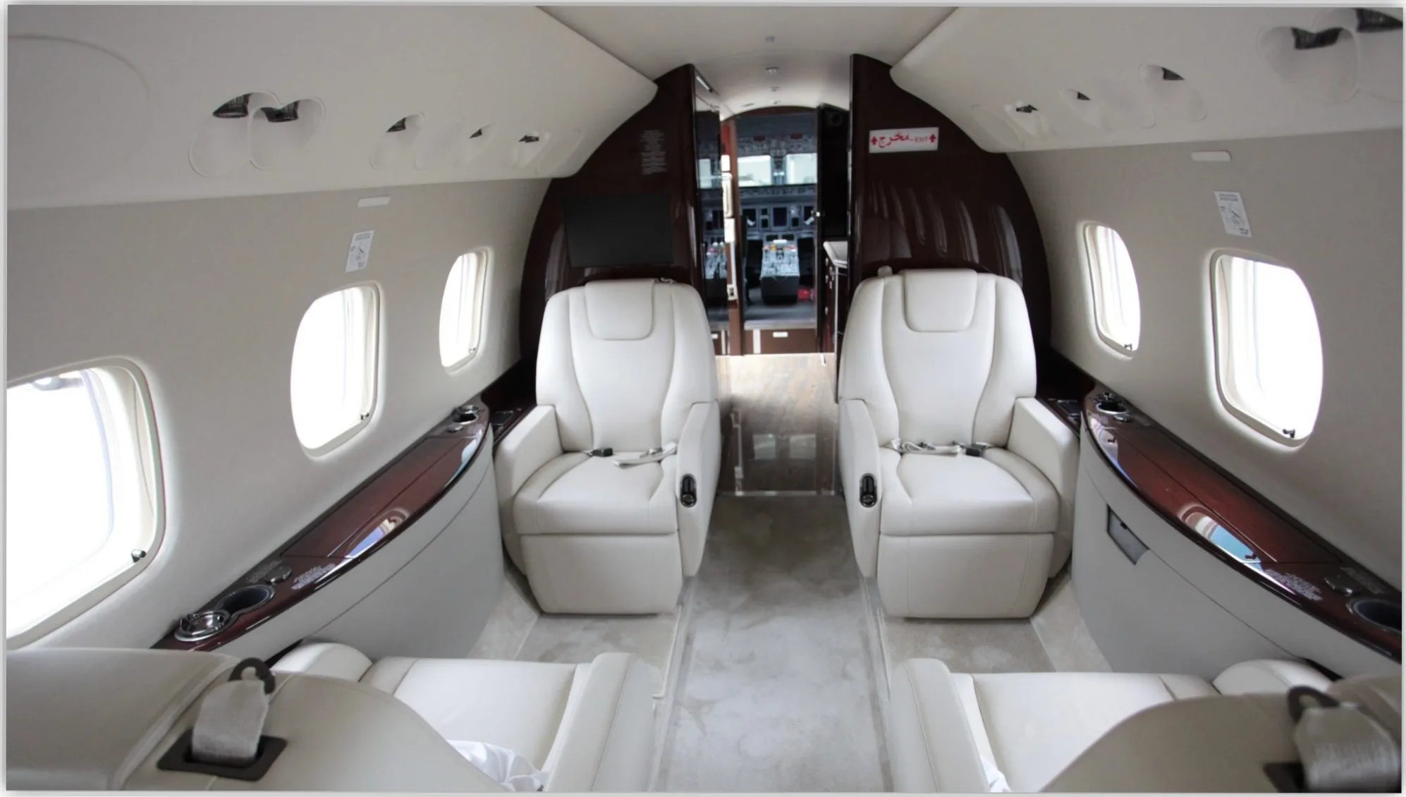 A view down the aisle of a private jet cabin, showing cream-colored seats, dark wood trim, and the open cockpit door in the distance.