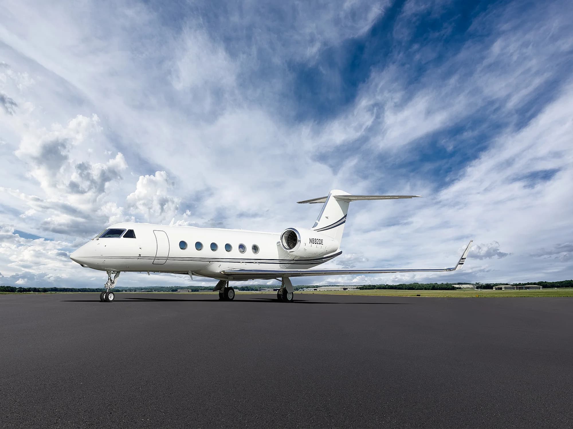 A white Gulfstream business jet with black and grey stripes on the tail is parked on a dark tarmac under a partly cloudy blue sky.