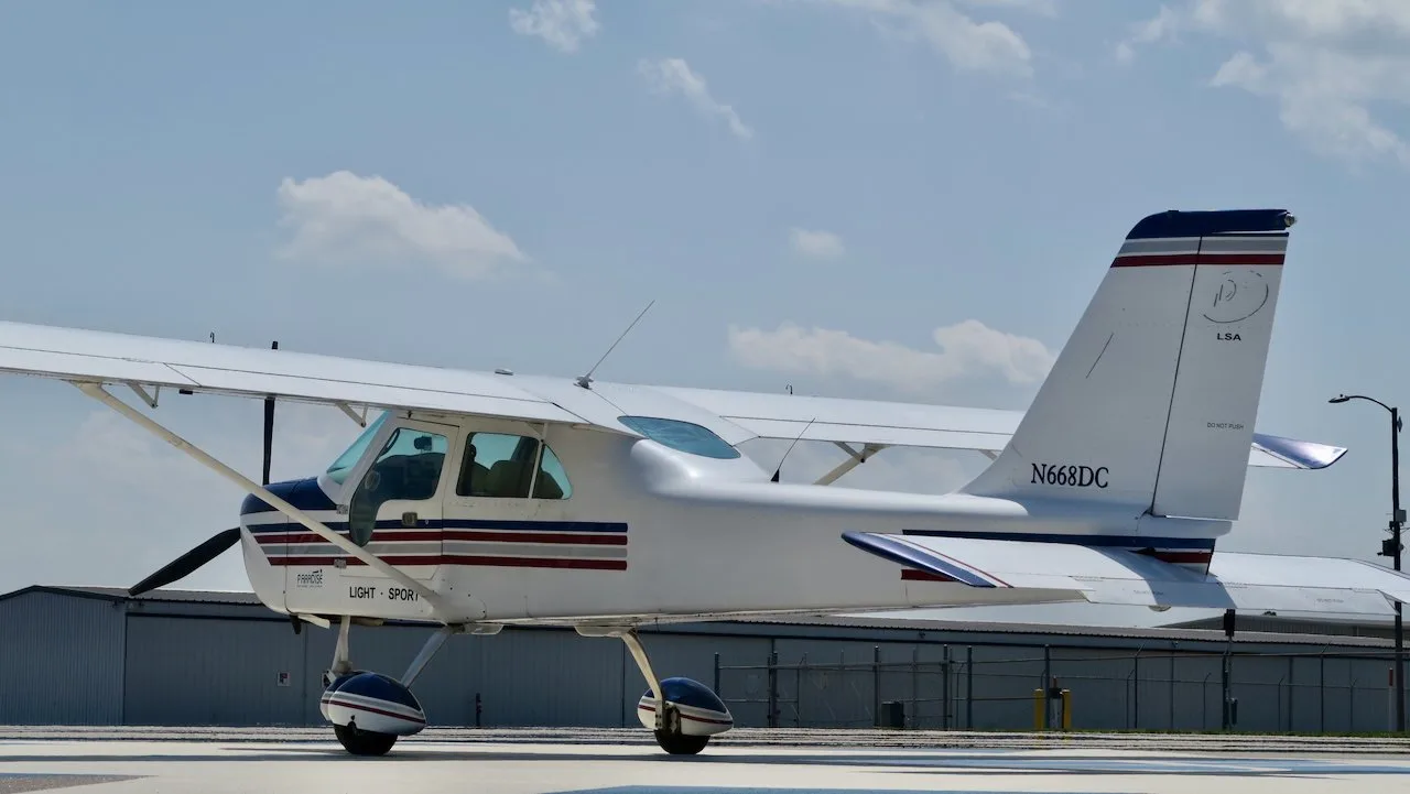 A rear-side view of a white Paradise P1 Light Sport aircraft with blue and red stripes, parked on a tarmac near hangars.