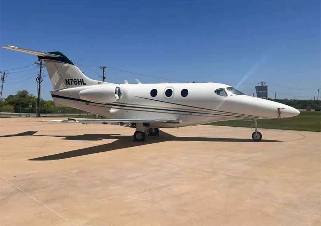 A white Beechcraft Premier 1 private jet, registration N76HL, with green and gold stripes, parked on a concrete tarmac under a clear blue sky.