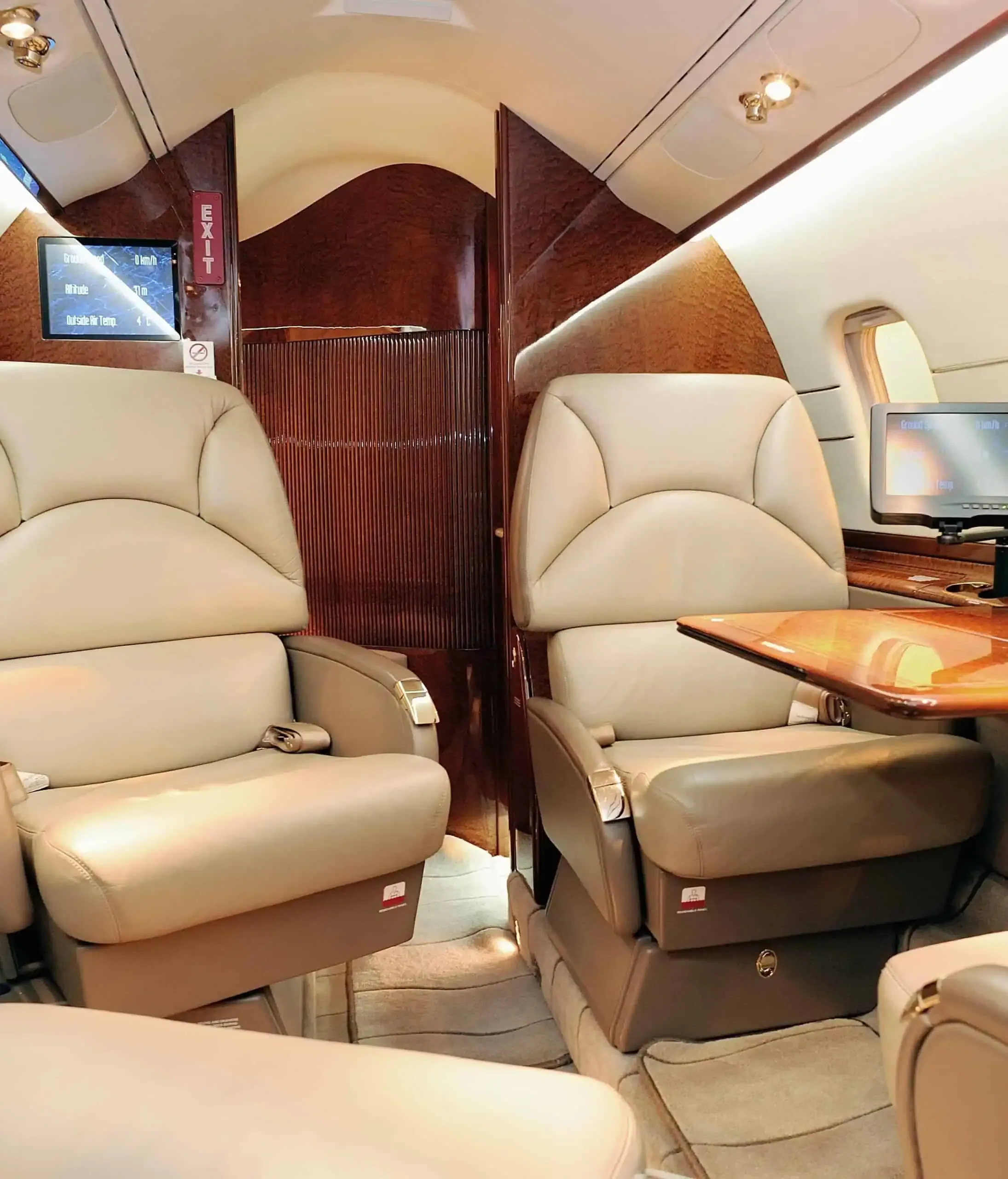 A luxurious aircraft cabin featuring two beige leather seats, a wooden table, and a monitor displaying flight information.