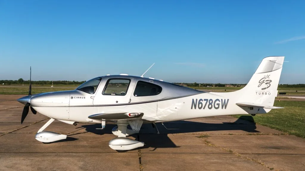 A side view of a silver and white Cirrus SR22 G3 Turbo aircraft (N678GW) on an airfield with a clear blue sky.