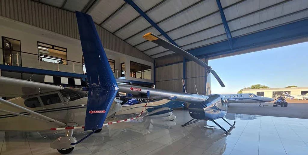 A low-angle view of a silver and blue Robinson R44 Raven II helicopter in a hangar, looking out towards the airfield.
