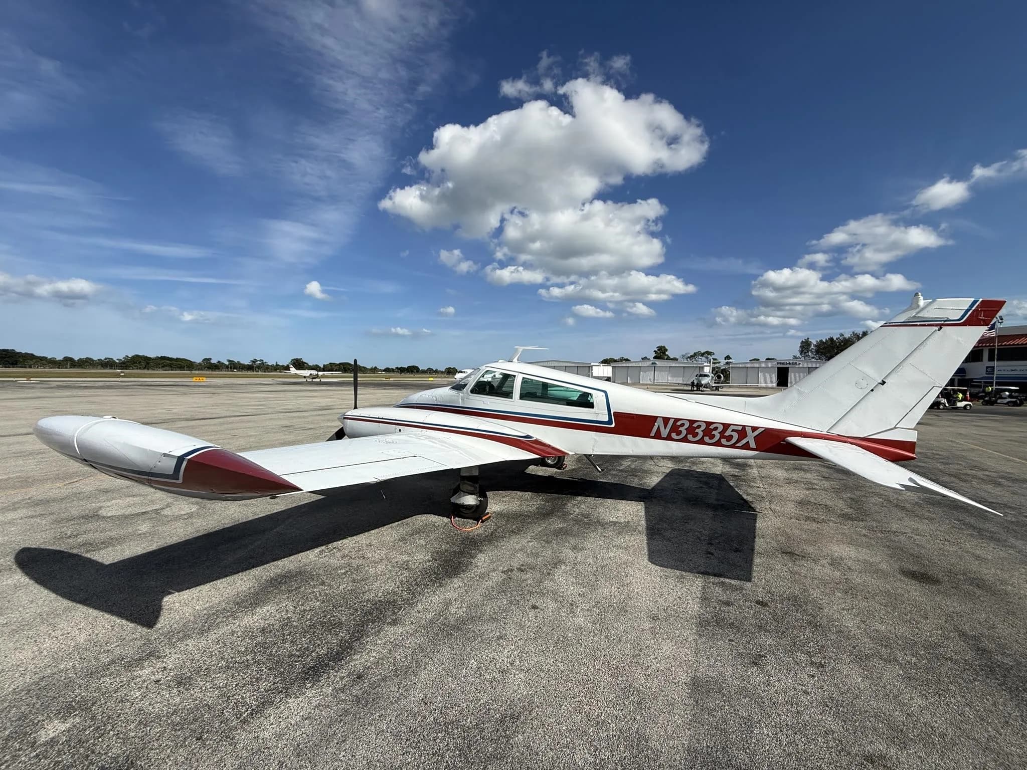 A white twin-engine propeller aircraft with red and blue stripes, registration N3335X, parked on an airport tarmac under a blue sky.