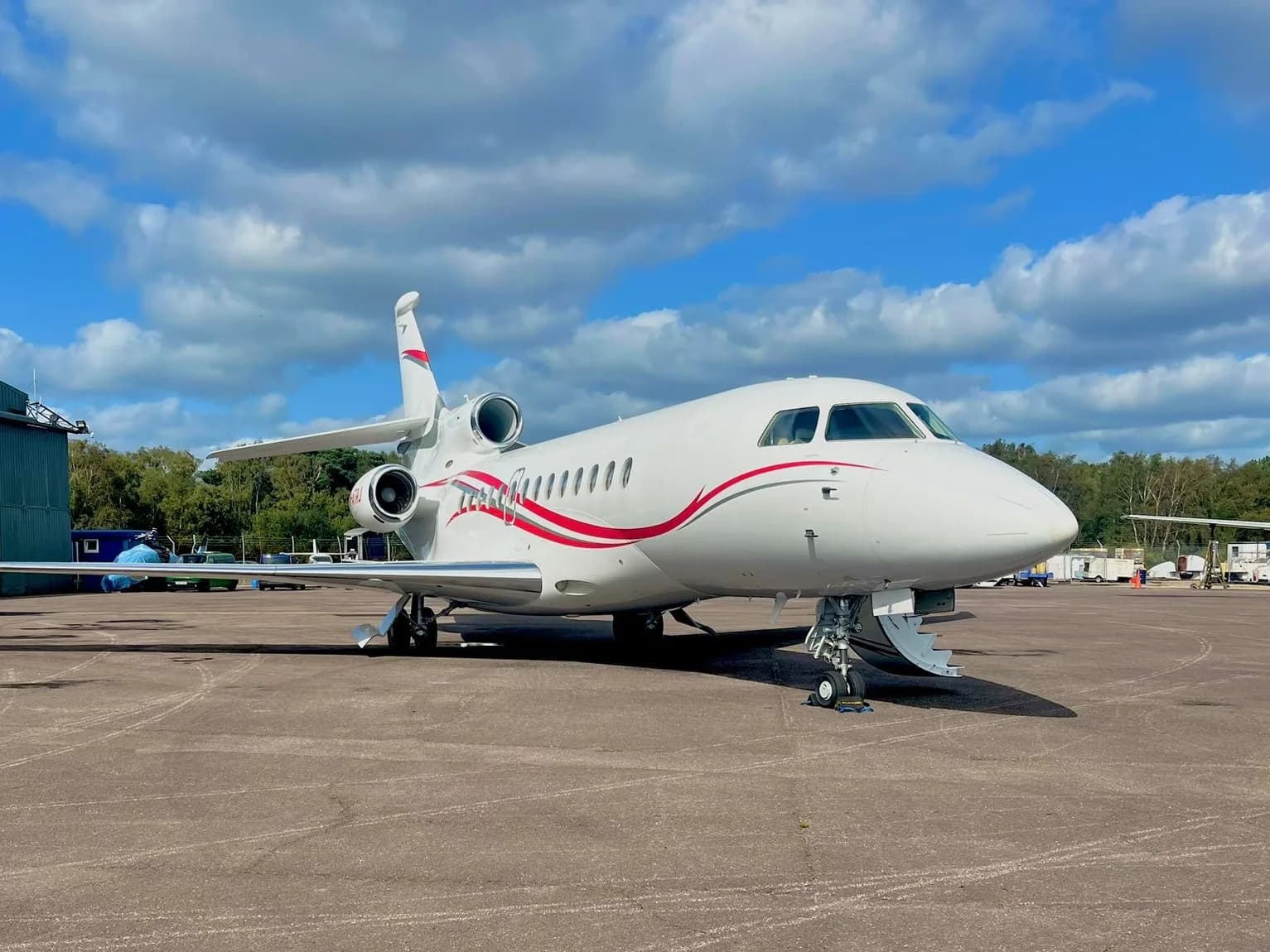 A white Dassault Falcon business jet with red and grey stripes is parked on an airfield under a partly cloudy sky.