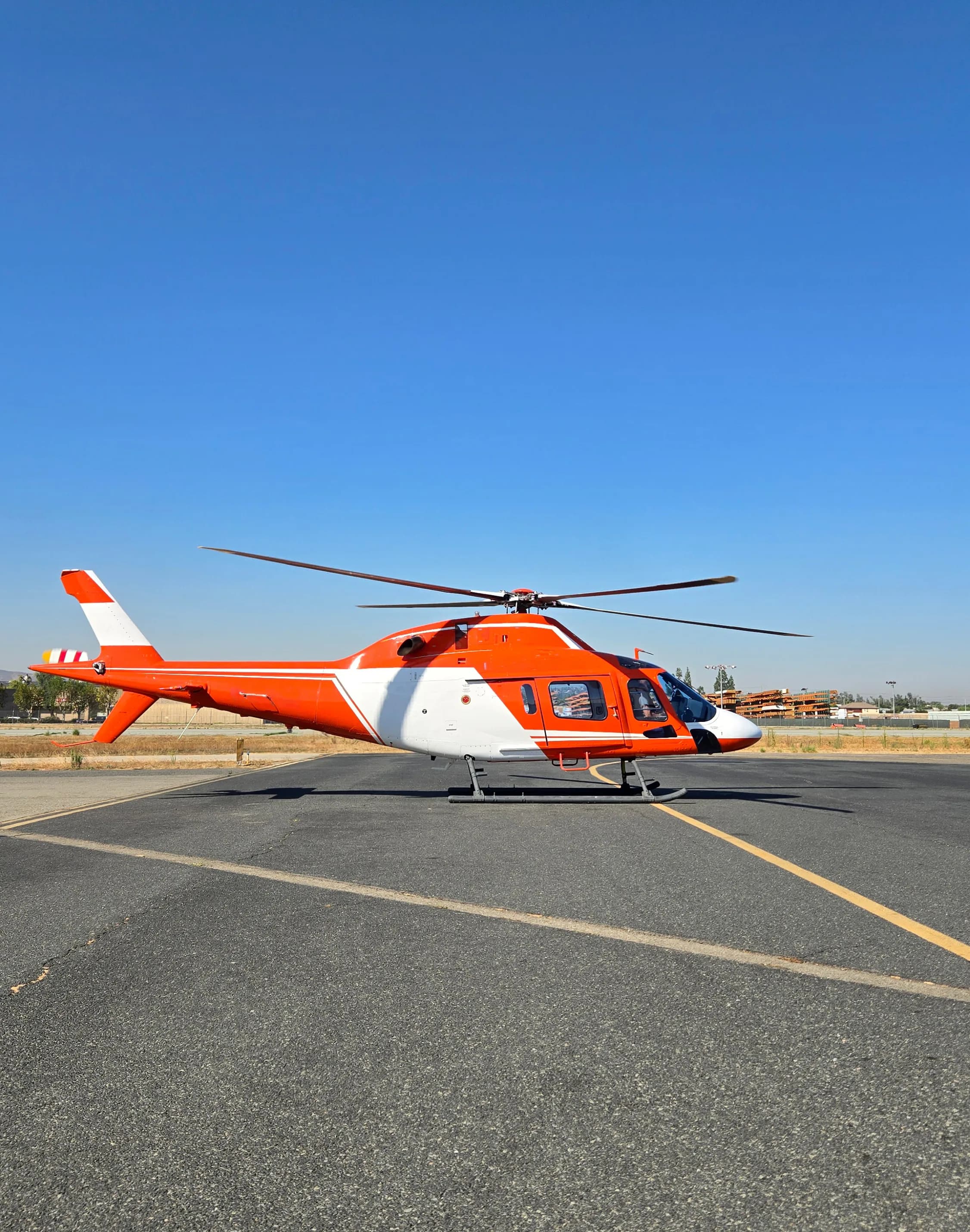 A red and white helicopter with a white nose and tail, parked on an asphalt runway under a clear blue sky.