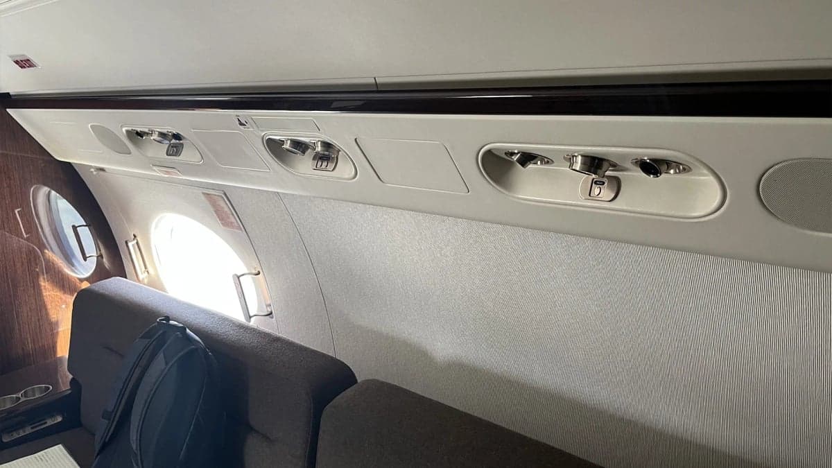 An interior view of an aircraft cabin, showing overhead panels with air vents and lights, and the top of two seats.
