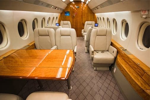 The spacious cabin interior of a private jet featuring cream leather seats and a large wooden table.