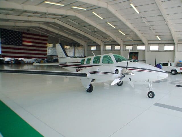 A white Beechcraft Baron N97R with red and black stripes is parked in a hangar with a large American flag in the background.