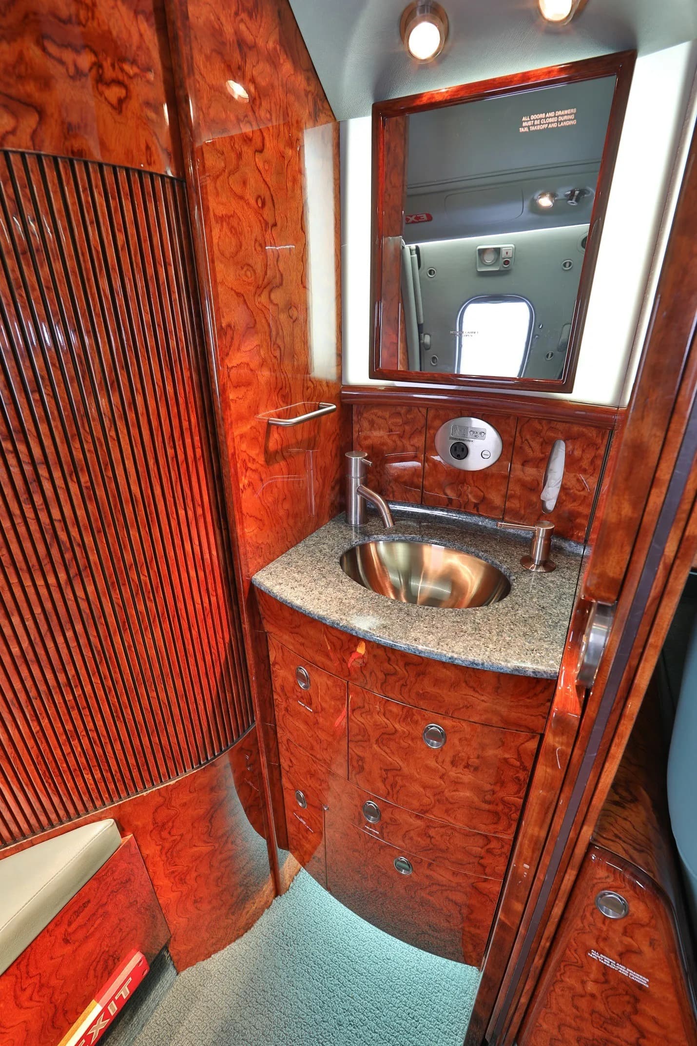 A private jet lavatory featuring a polished wood vanity with a metal sink, mirror, and storage.