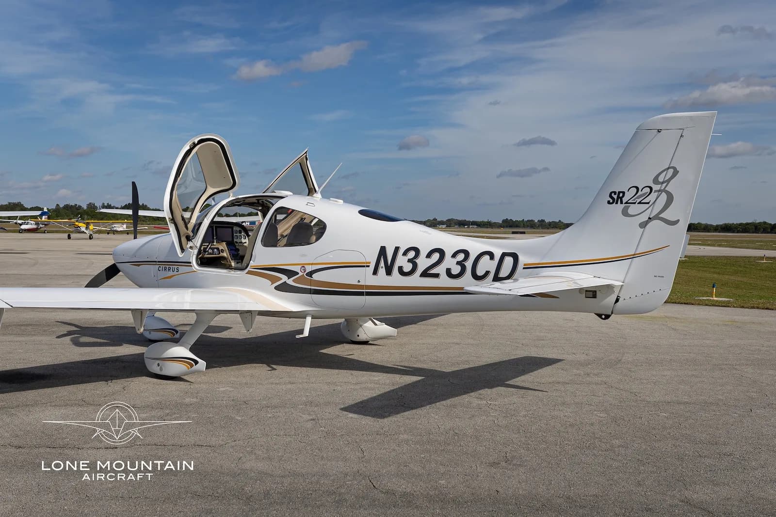 A white Cirrus SR22 aircraft with gold and black accents, registration N323CD, parked on an airfield with its doors open, viewed from the right side.