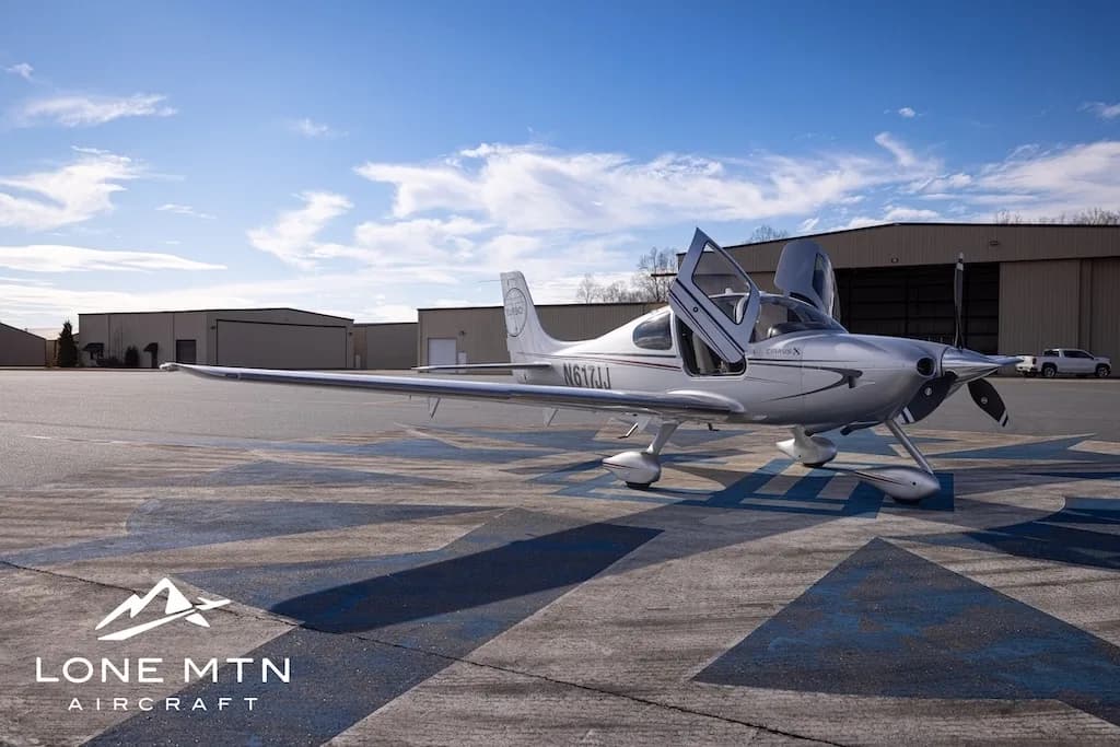 A silver Cirrus SR22T aircraft, N617JJ, with its doors open, parked on an airfield with hangars in the distance.