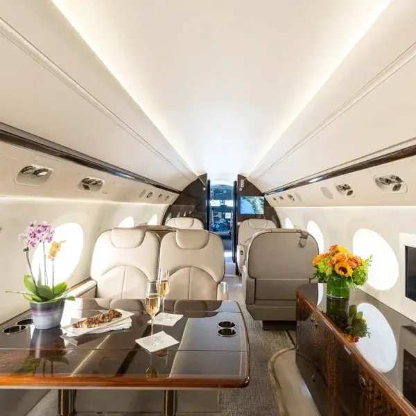 A luxurious aircraft cabin interior with light-colored leather seats, a polished wooden table set with champagne and pastries, and floral arrangements.
