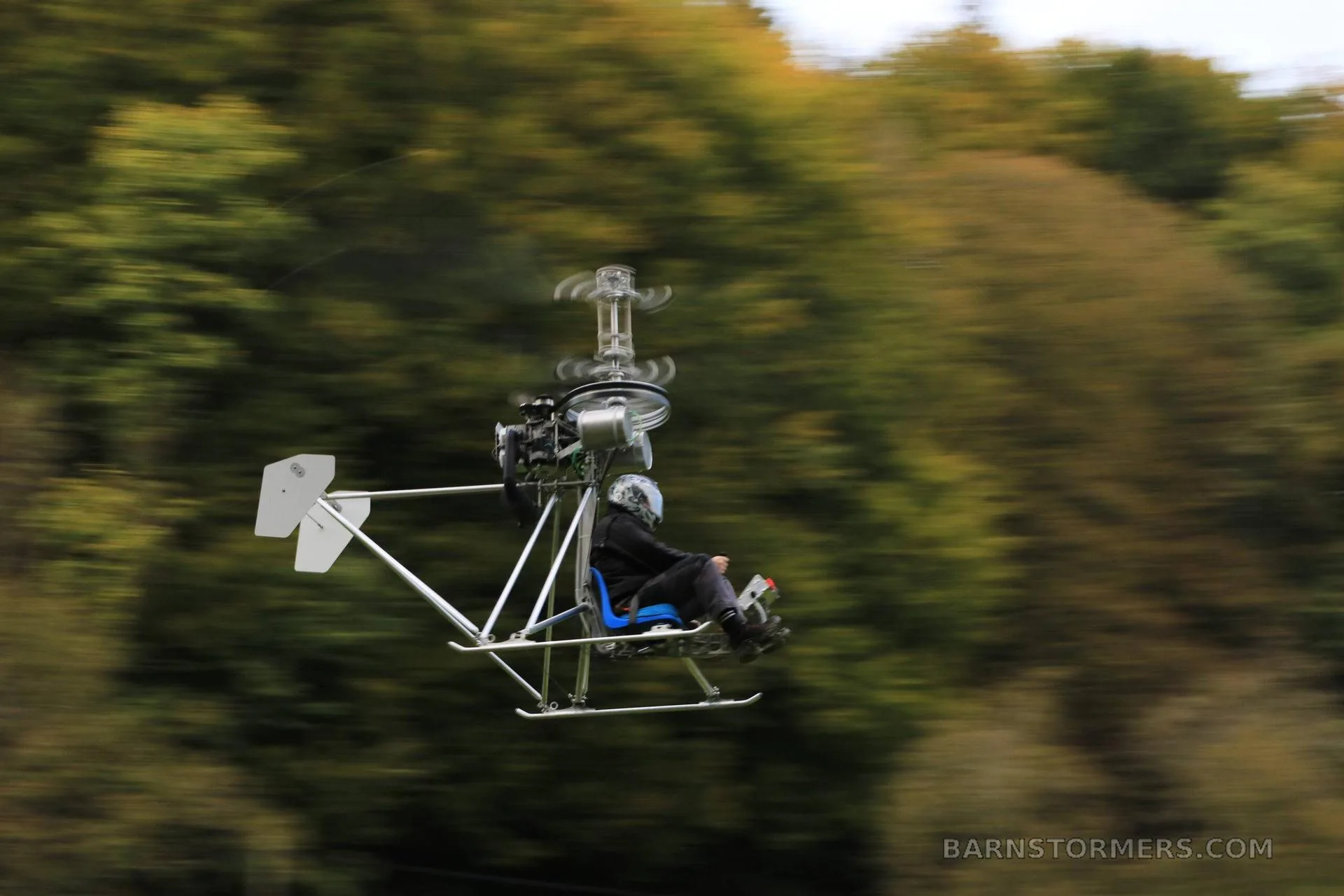 A single-seat ultralight helicopter with a pilot in a helmet flies past a blurred green and brown treeline.