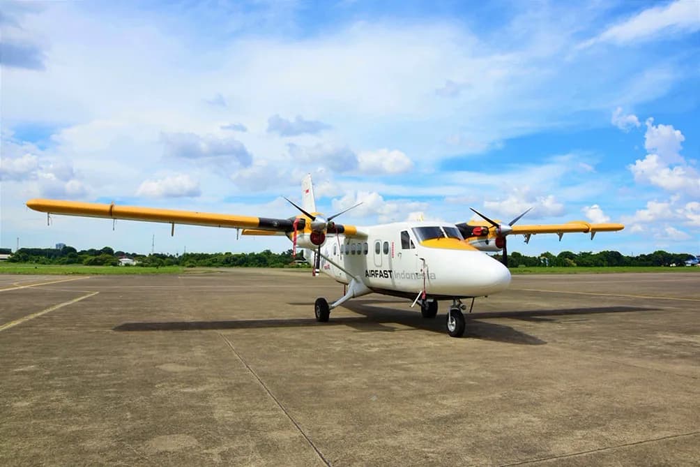 A white and yellow Airfast Indonesia Twin Otter aircraft parked on an airfield under a partly cloudy sky.