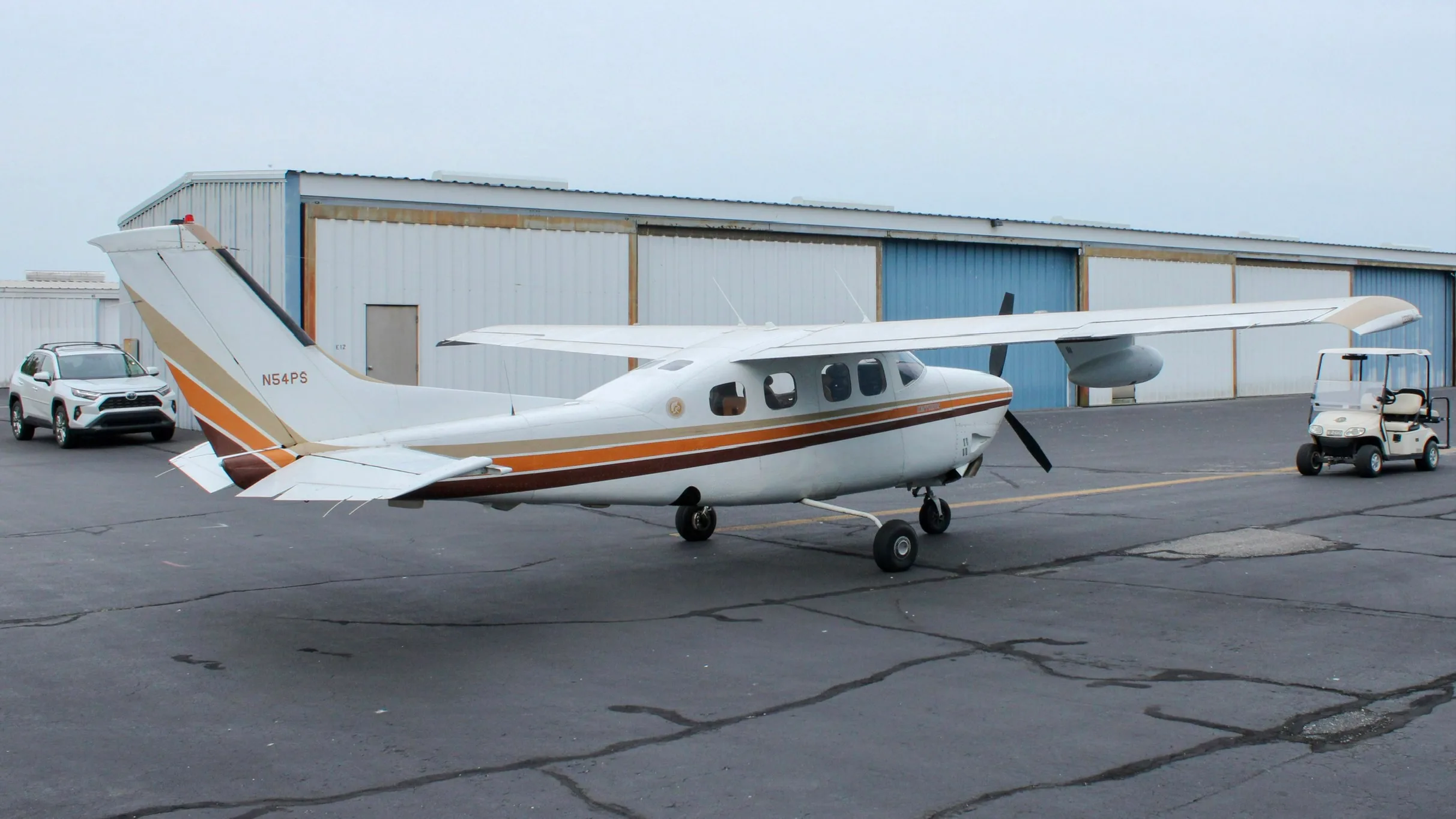 A white Cessna P210N Centurion, registration N54PS, parked on an airport apron with hangars and a golf cart in the background.