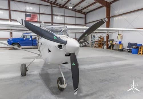 A close-up front view of a white light sport aircraft with a two-blade propeller, parked in a hangar.
