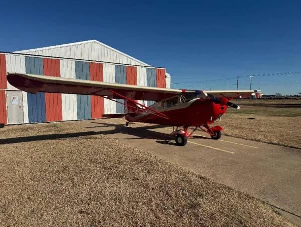 A red and cream high-wing taildragger aircraft parked on an airfield next to a striped hangar.