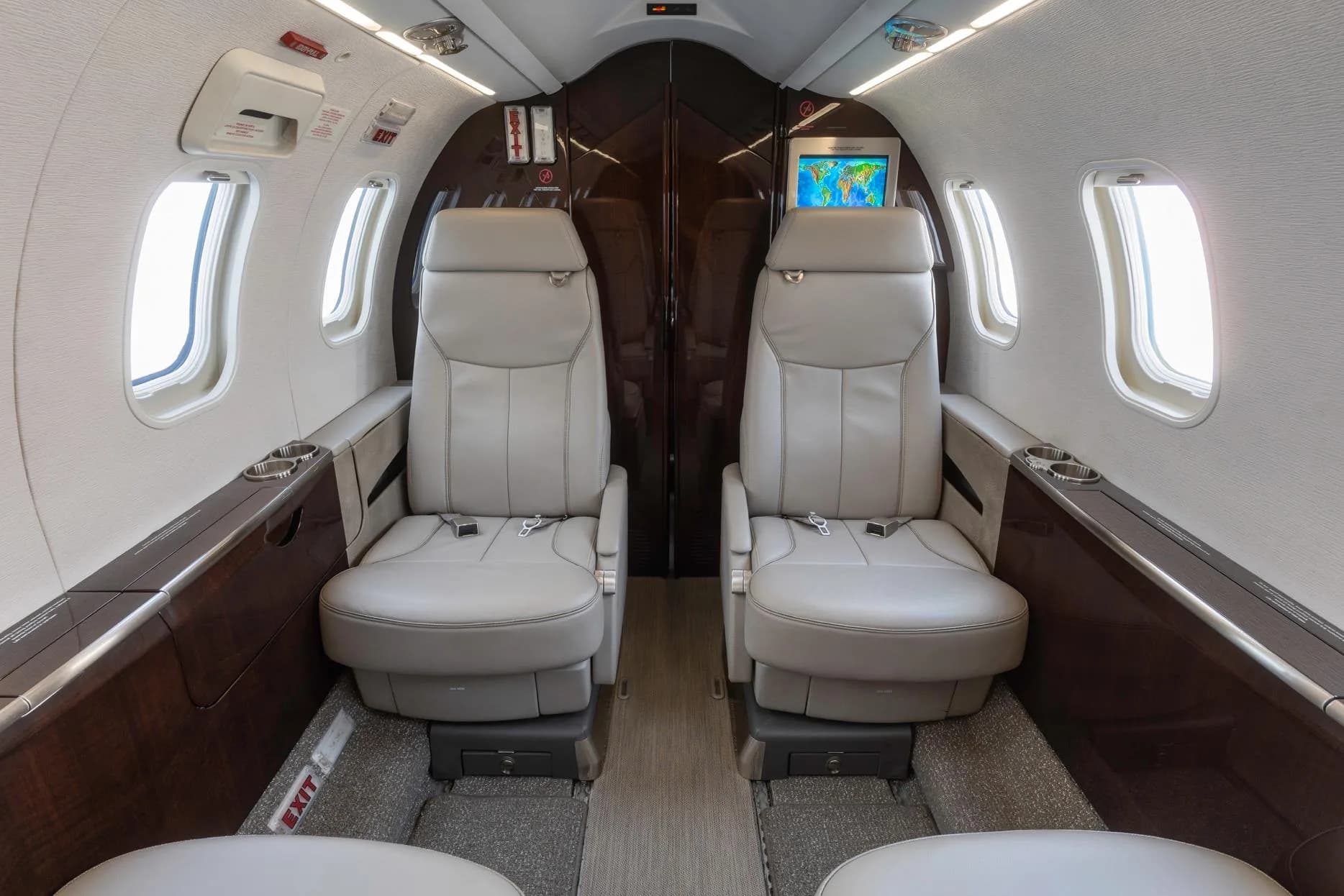 View of two light gray leather seats in a private jet cabin, with a monitor displaying a map at the rear.