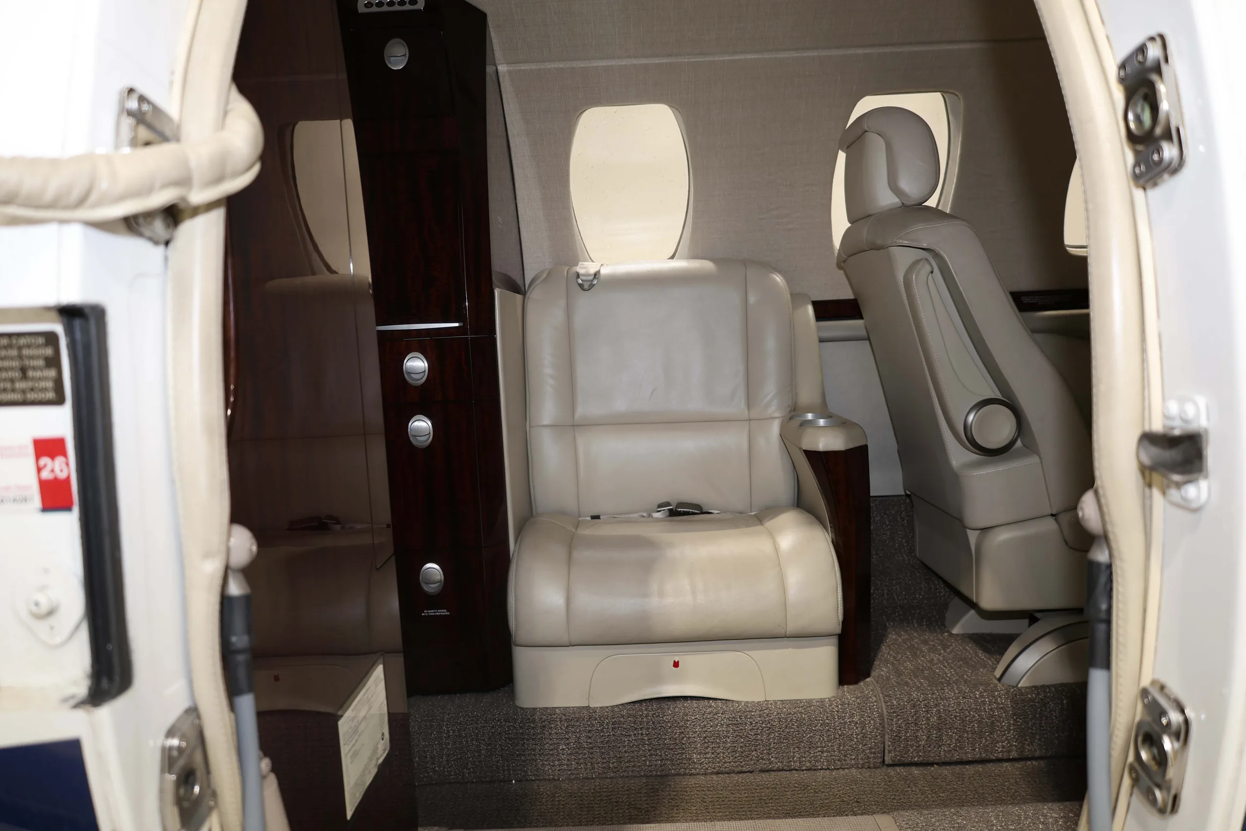 The interior cabin of a business jet viewed from the open main door, showing light beige seats and a dark wood cabinet.