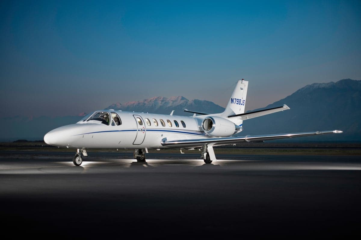 A white Cessna Citation business jet with blue stripes is parked on a tarmac at dusk, with mountains in the background.