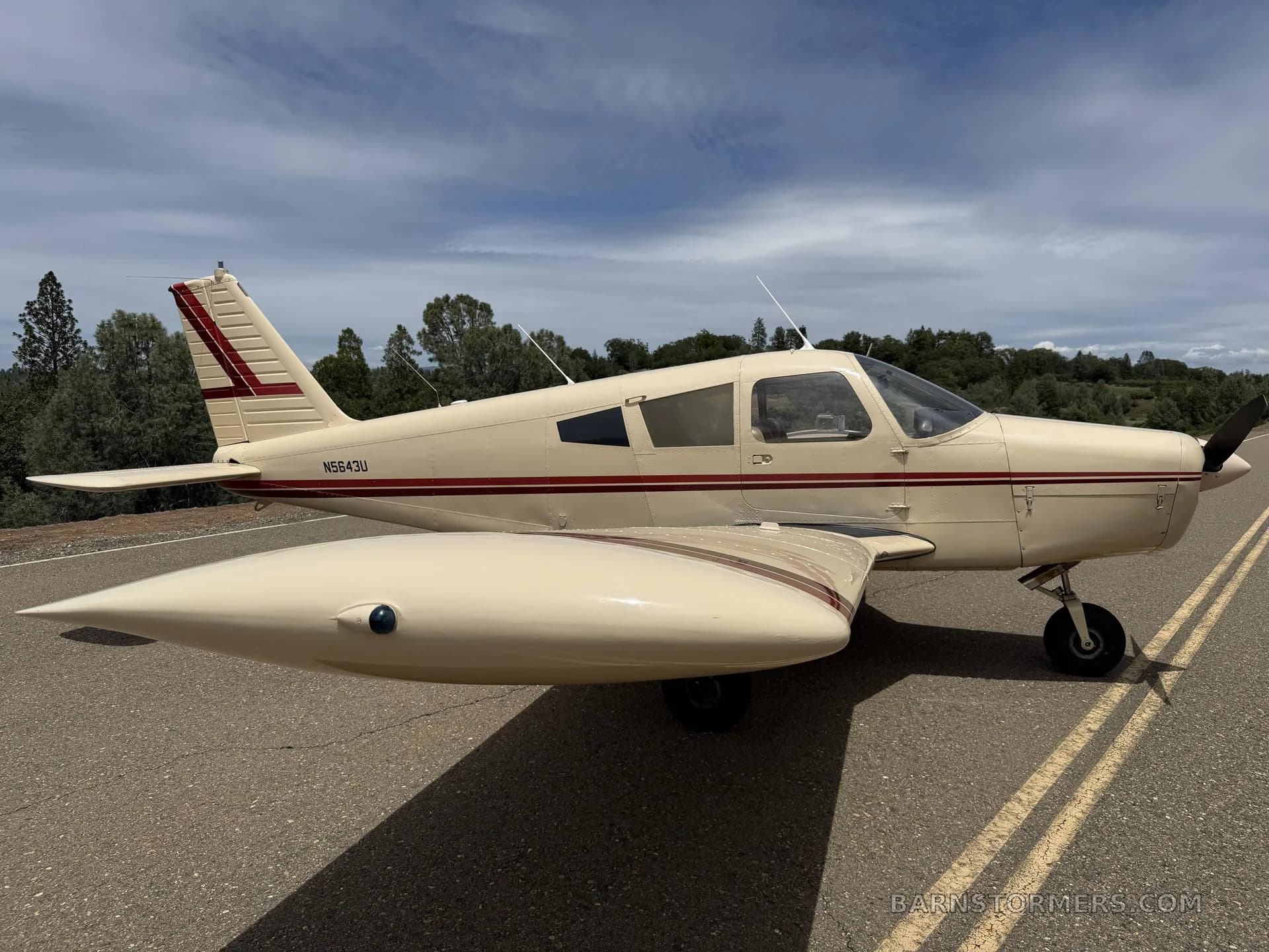 A cream-colored Piper Cherokee with red stripes, registration N5643U, parked on an asphalt surface under a partly cloudy sky, viewed from the front-right side.