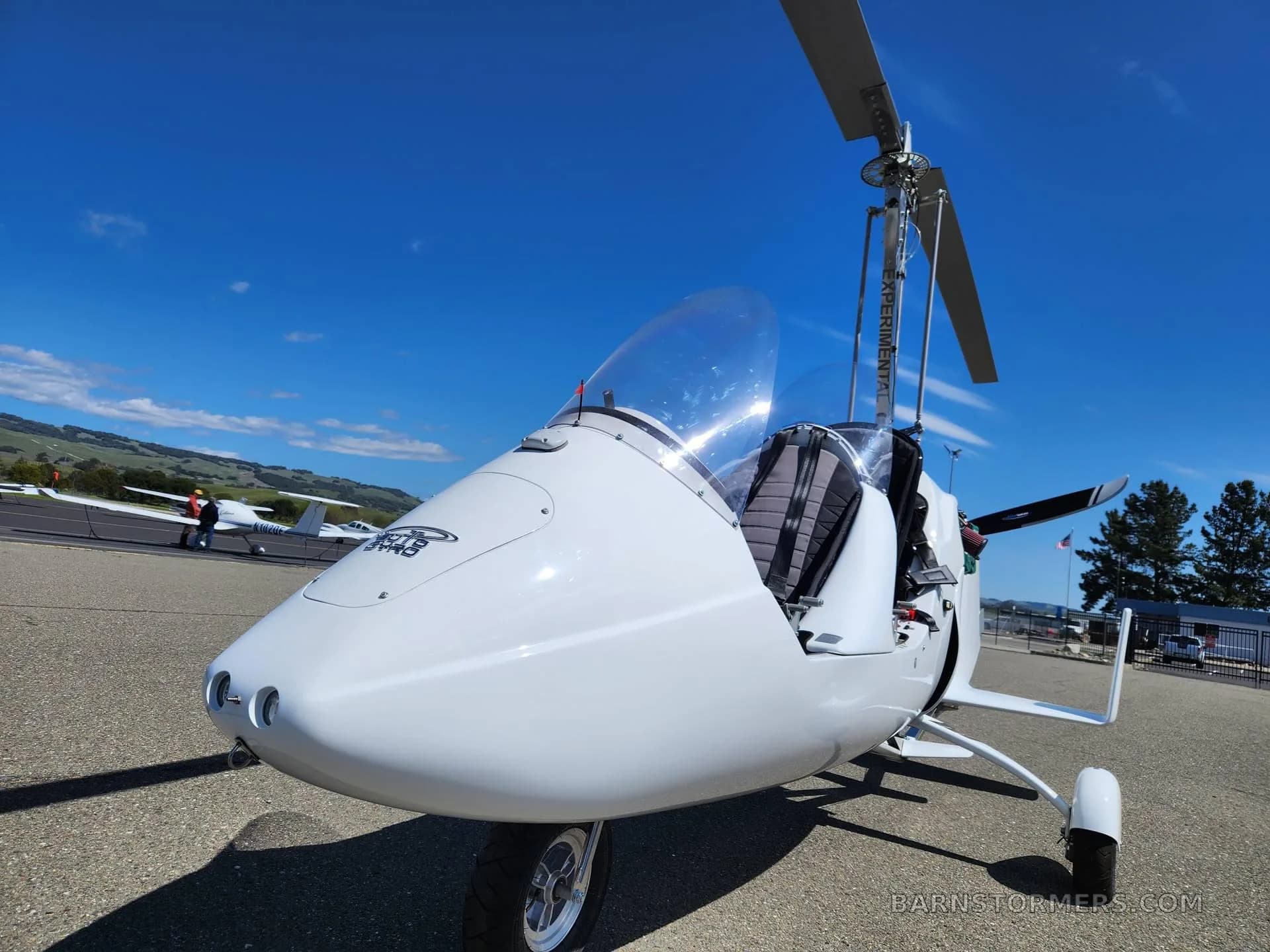 A close-up front view of a white gyrocopter with a clear bubble canopy, parked on an airfield.