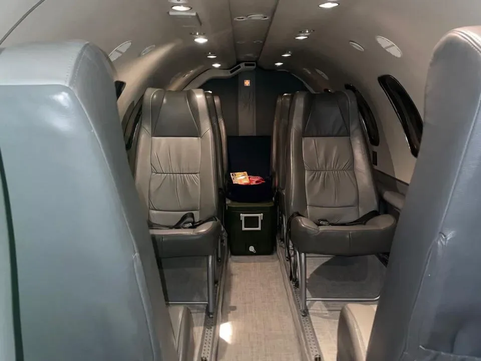 A view of the aircraft interior cabin, showing grey leather seats and a cooler box in the aisle.