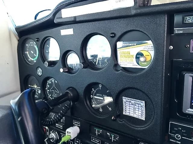 A close-up view of an aircraft's instrument panel, showing various gauges, a carbon monoxide detector, and a fuel calibration chart.