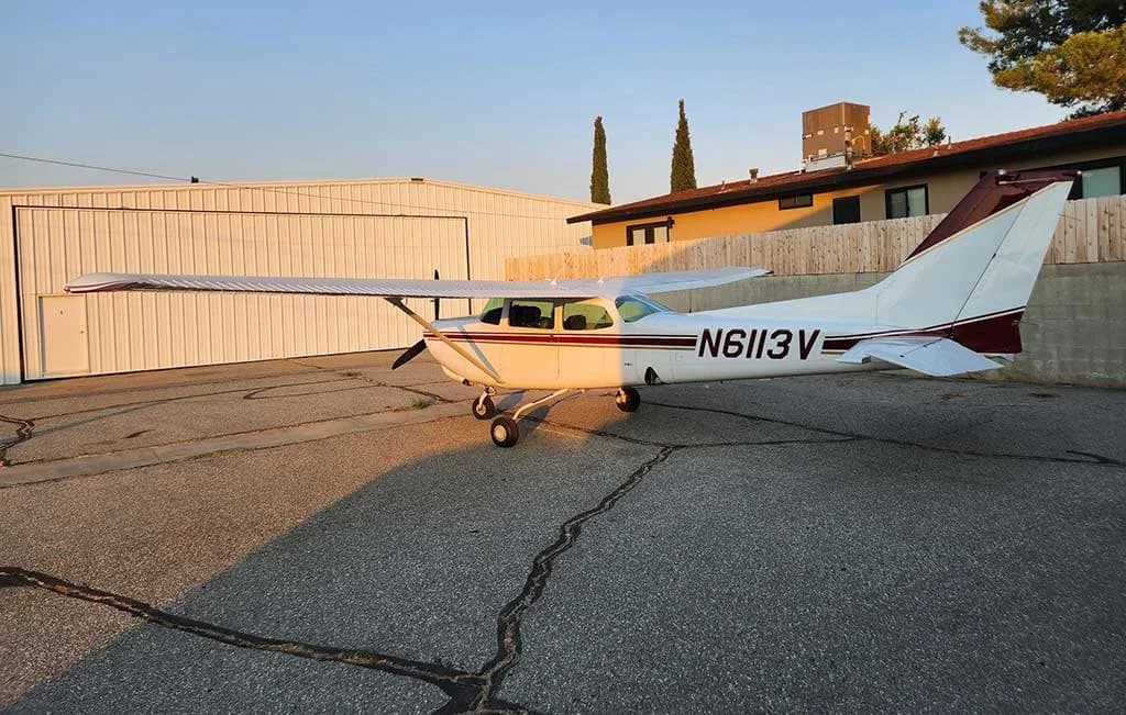 A side view of a white and maroon Cessna 172 aircraft, N6113V, parked on an asphalt apron next to a hangar.