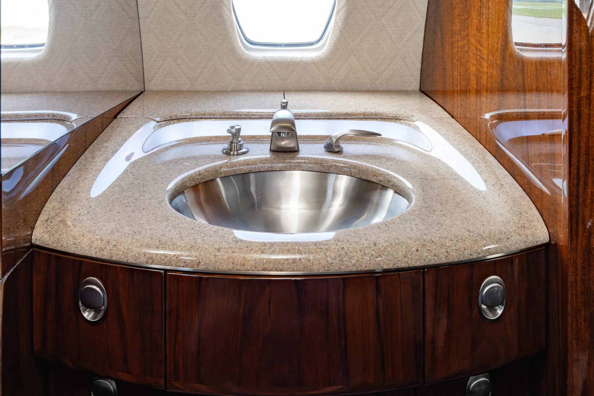 A polished metal sink set into a speckled countertop with dark wood cabinetry in an aircraft lavatory.