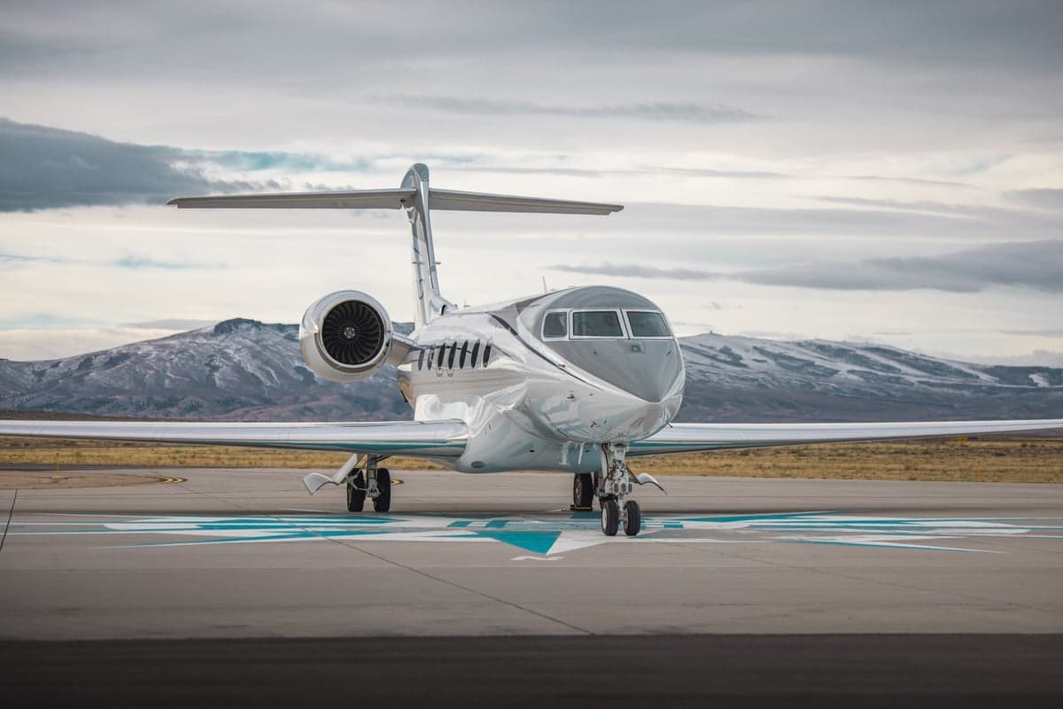 A sleek silver business jet with dark accents parked on an airfield with snow-capped mountains in the background.