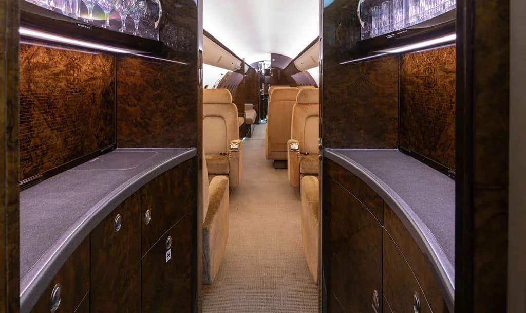 A view from the galley of a private jet looking into the main cabin, showing dark wood cabinetry and plush tan seats.