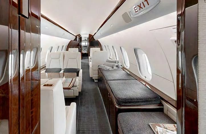 The interior of a private jet cabin with cream seats, a grey divan, and dark wood finishes.