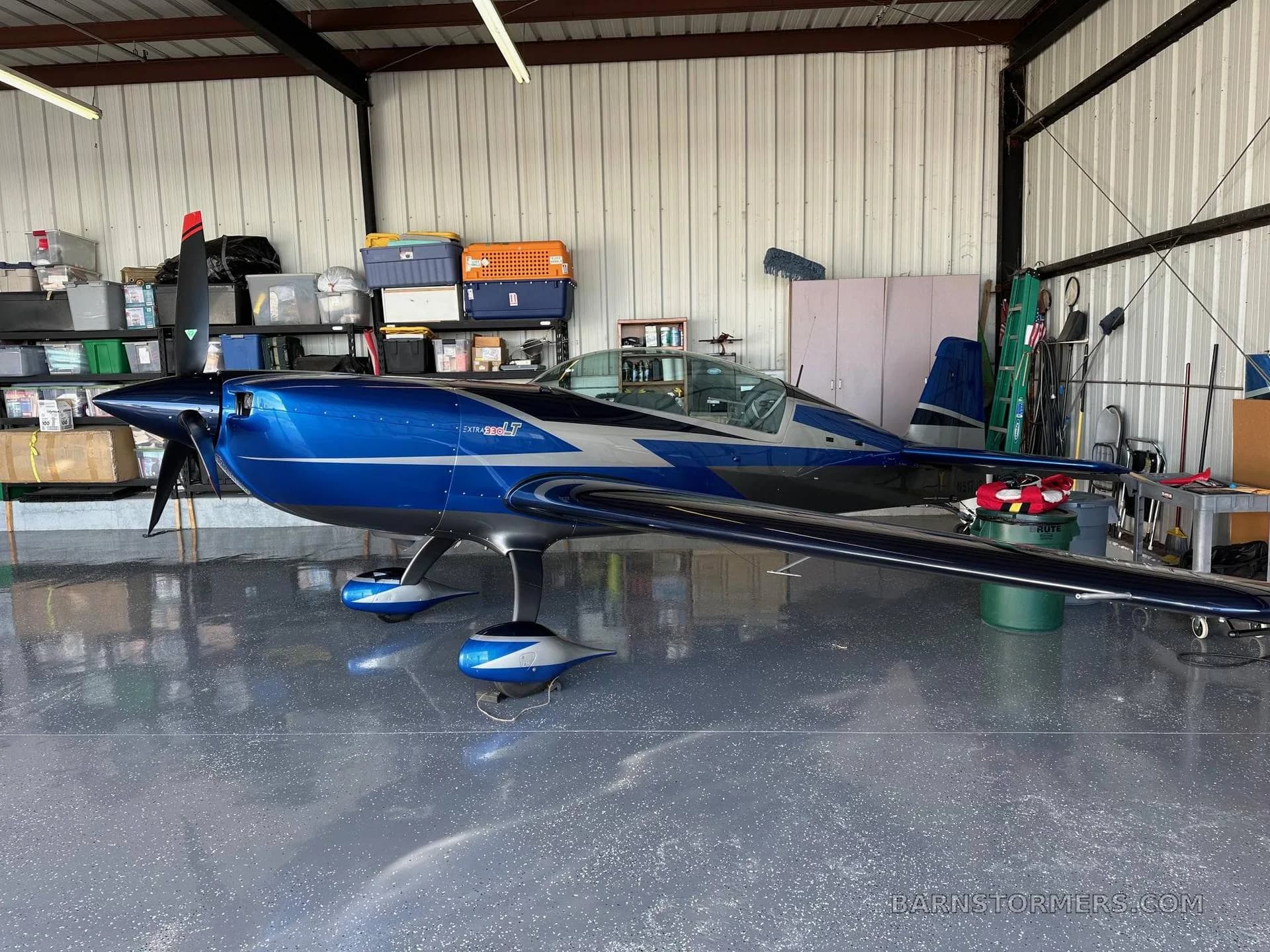 A blue and silver Extra 300LT aerobatic aircraft is parked inside a hangar with various items stored in the background.