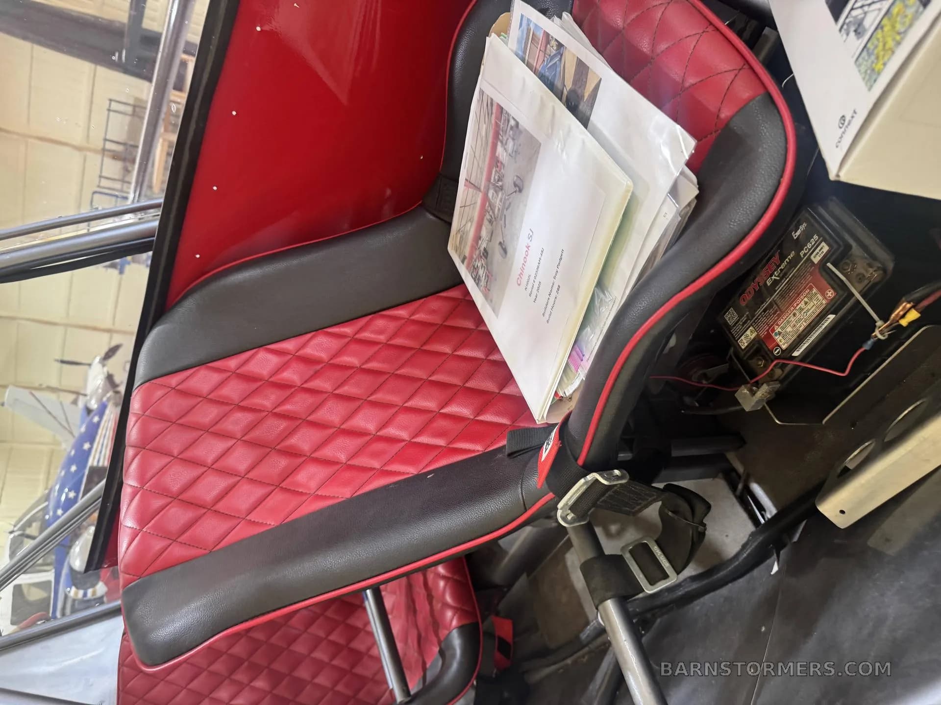 A red and black quilted aircraft seat with a binder of documents resting on it, seen from an interior perspective.