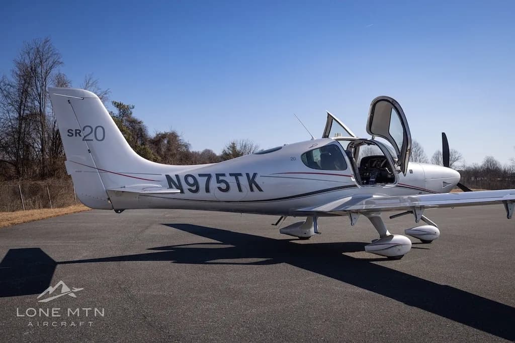 A silver Cirrus SR20 aircraft with registration N975TK parked on an asphalt runway, viewed from the rear-right with doors open.